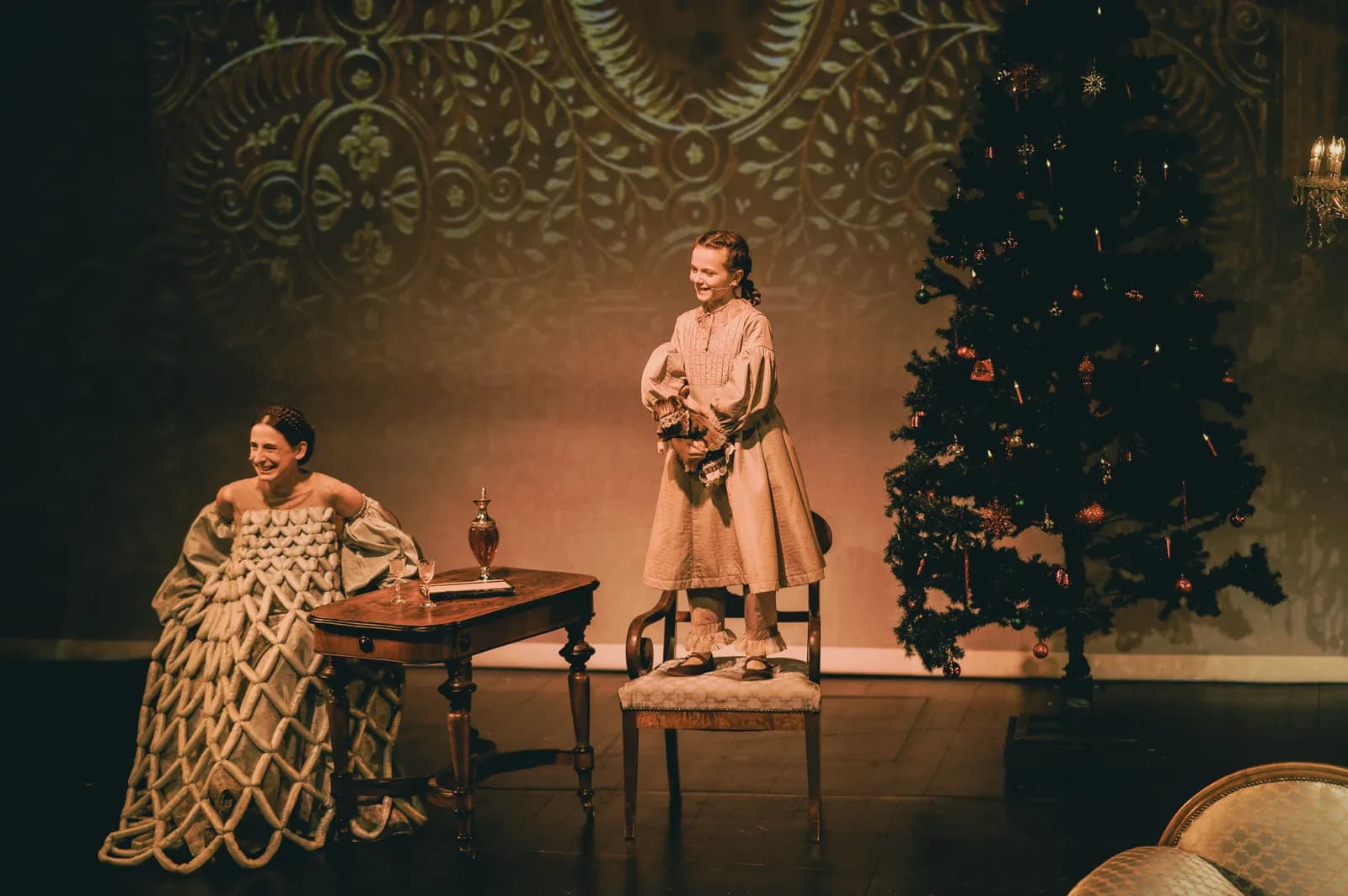 Na zdjęciu scena teatralna z dwiema osobami w historycznych kostiumach. Jedna siedzi przy stoliku (Katarzyna Michalska), druga (Zofia Habasińska) stoi na krześle obok. W tle choinka i dekoracyjna ściana.