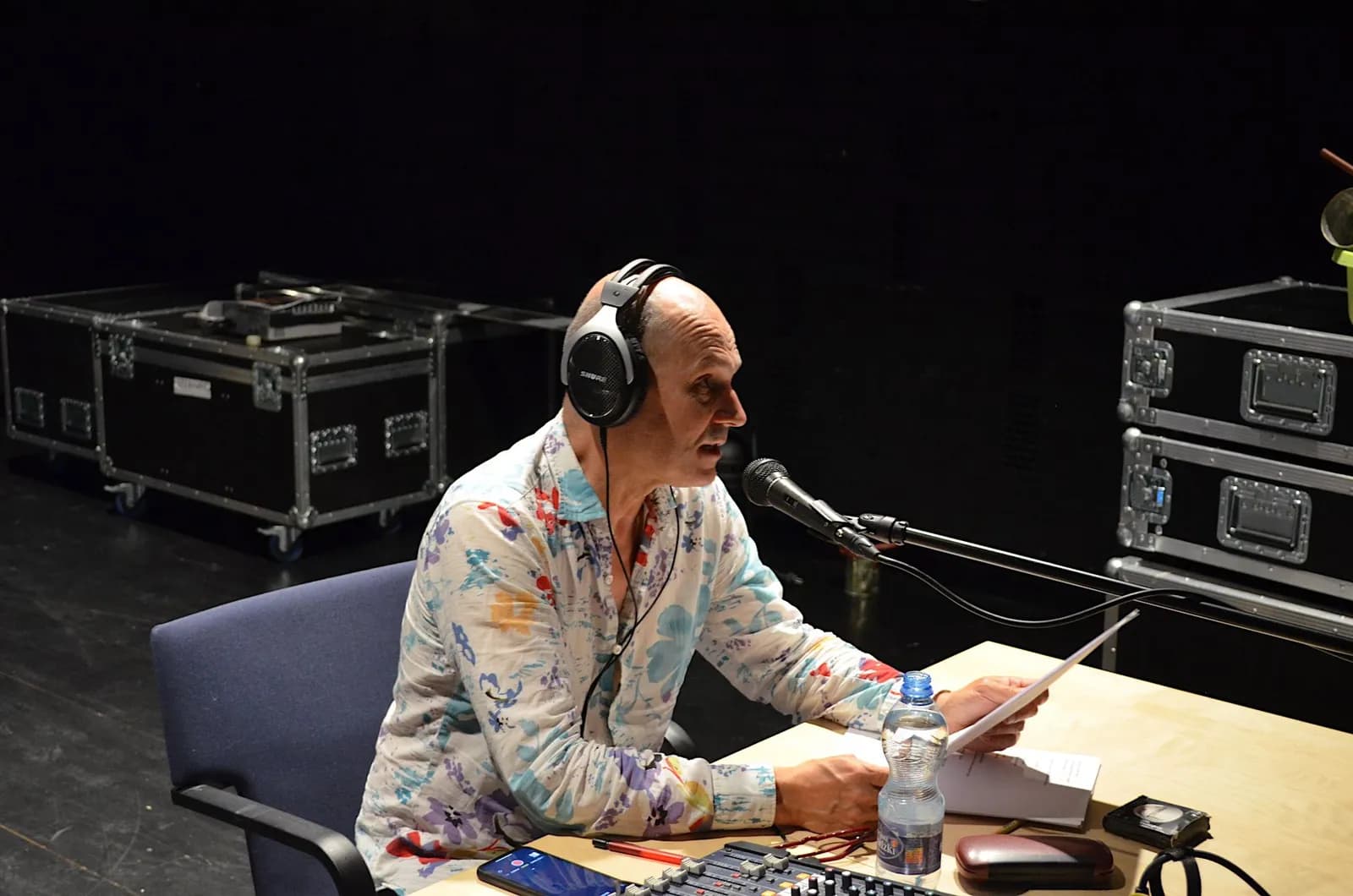 Lektor audiodeskrypcji w trakcie odczytu. Jest łysy, ma kwiecistą koszulę. Na uszach słuchawki, przed nim mikrofon, czyta z kartki.